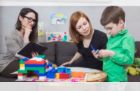 Prostokątna grafika ze zdjęciem na którym pokazane jest dziecko bawiące sie klockami w towarzystwie dwóch siedzących koiet - matki i psychologa.