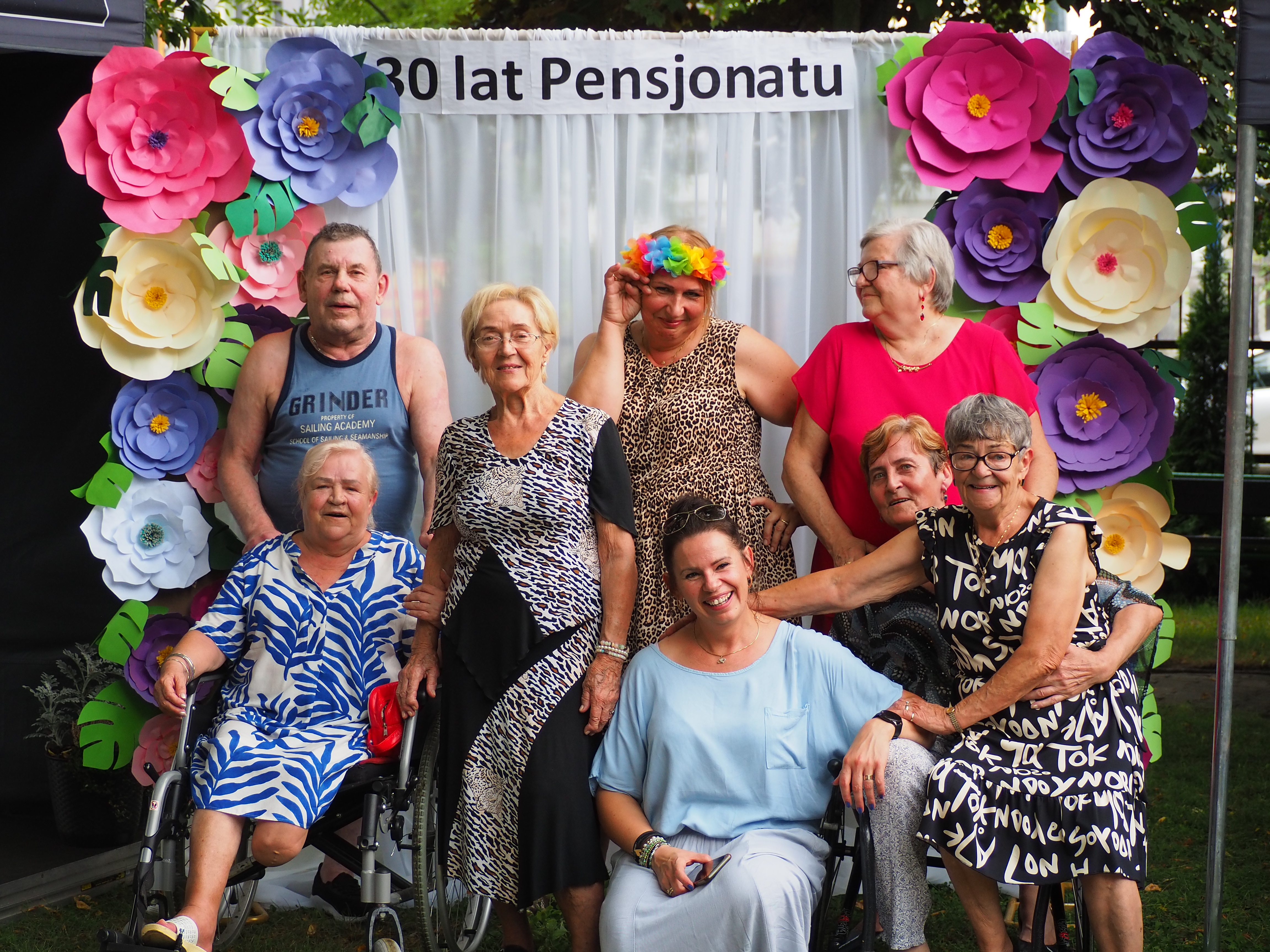 9 osób w kolorowych ubraniach pozuje na tle ozdobnego napisu 30 lat pensjonatu oraz sztucznych kwiatow.