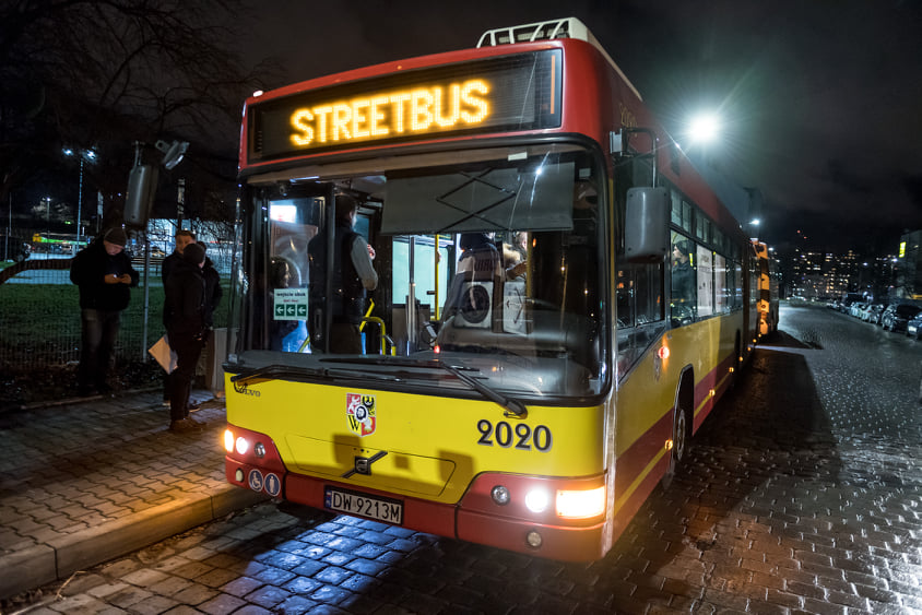 Na zdjęciu jest noc. Na pierwszym planie stoi żółto-czerwony miejski autobus ze świecącym napisem Streetbus