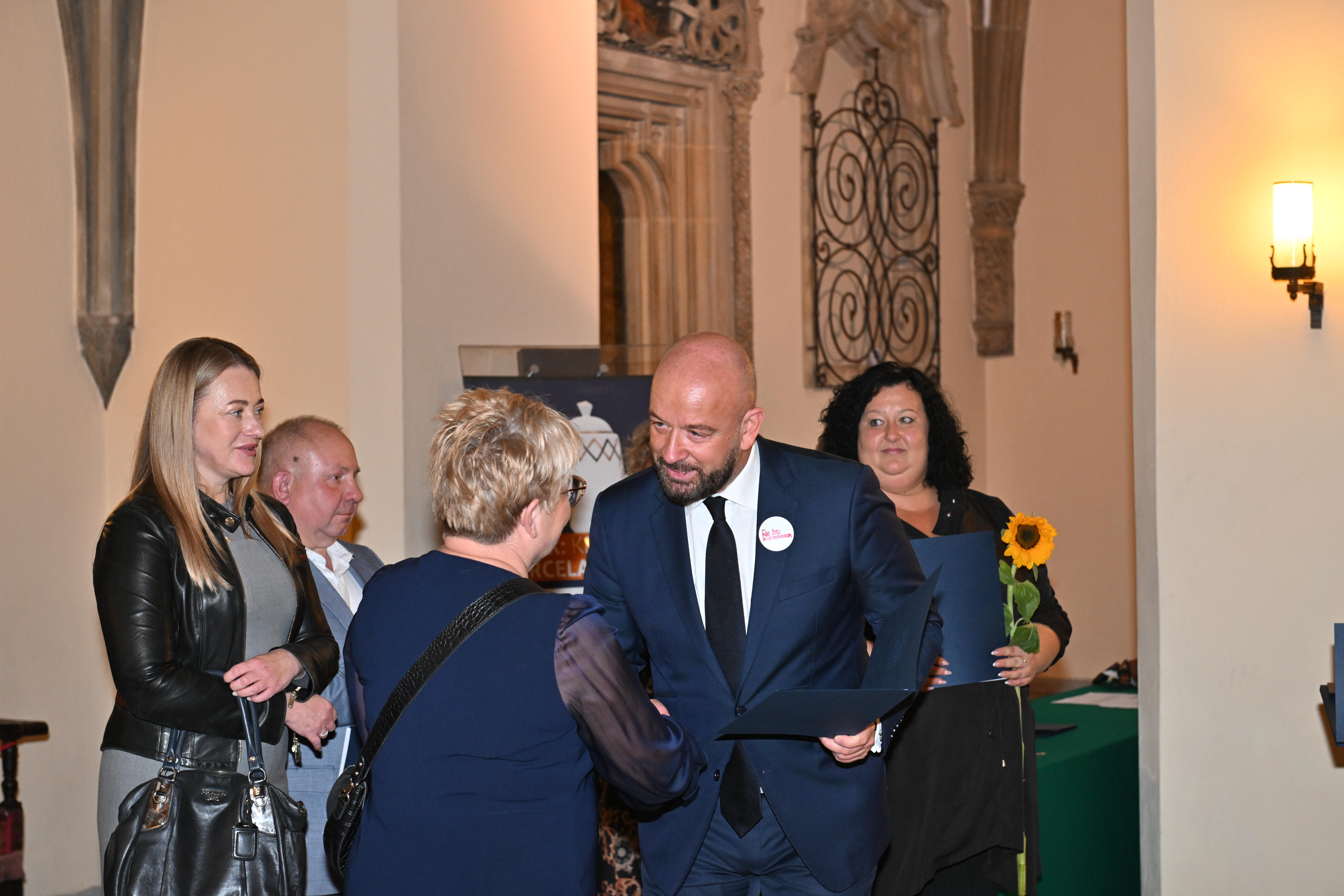 Zdjęcie przedstawia Spotkanie z opiekunkami i opiekunami zorganizowano w Sali Wielkiej Ratusza we Wrocławiu. Grupa opiekunek w szeregu przyjmuję podziękowania przez uścisk dłoni od  Panie Prezydenta Jacka Sutryka. Panie trzymają kwiat – słonecznika. 